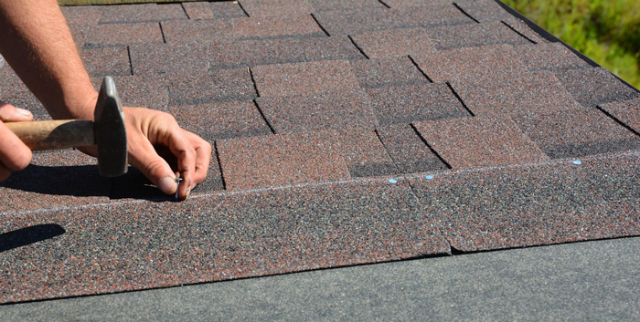 Roofer hands installing asphalt shingles on house construction roof corner with hammer and nails in motion.